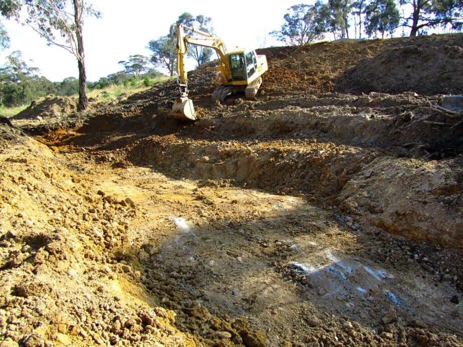 Excavator building a dam in a rural area with trees in the background.