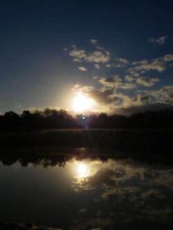 Sunset over a calm water body, reflecting the sky and horizon, with a Big Ditch dam construction site nearby.