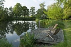 Tranquil lakeside scene with wooden deck and chairs in lush greenery.