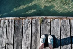 Weathered wooden dock over clear water, feet in sneakers, serene waterfront scene, outdoor leisure.